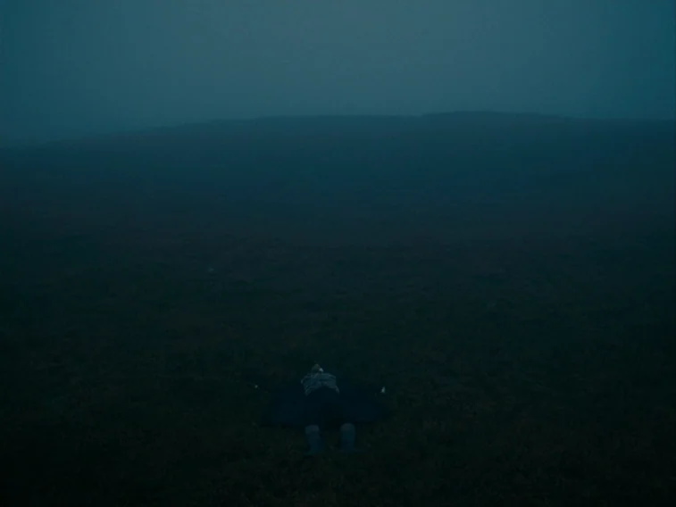Wuthering Heights movie still: wide — A vast, dark, fog-shrouded moorland stretches out under a heavy, overcast sky. A single figure lies motionless…