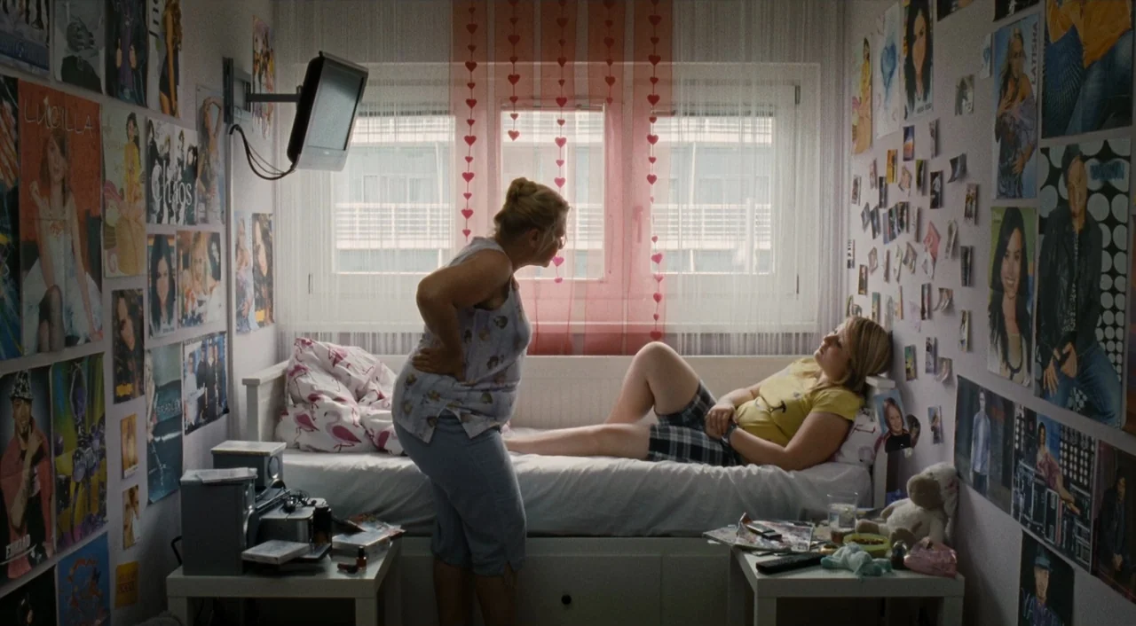 Paradise: Love (2012) movie still: medium — A small bedroom features walls completely covered in posters and photos. An older woman stands over a…