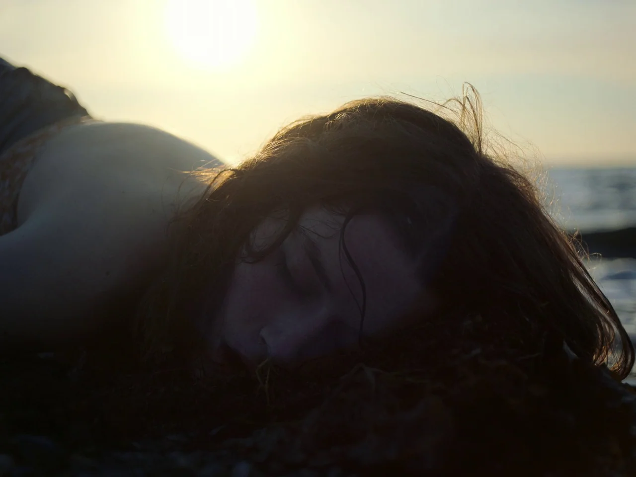 Of An Age (2022) movie still: closeup — A shoreline with dark wet seaweed scattered on the sand at sunset. A young woman lies face-down on the…