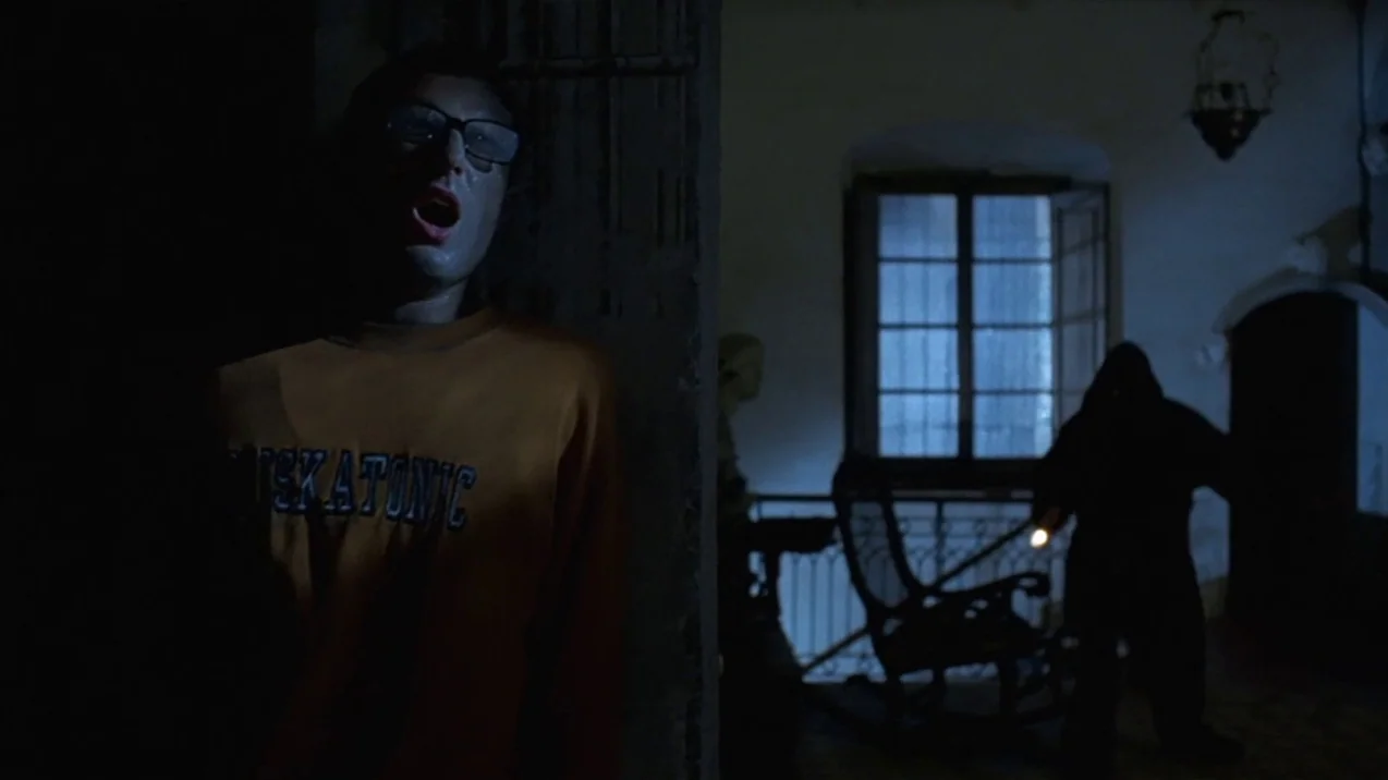 Dagon (2001) movie still: medium — A dark, shadowed hallway features a wooden rocking chair and a window with moonlight. A man in a yellow…