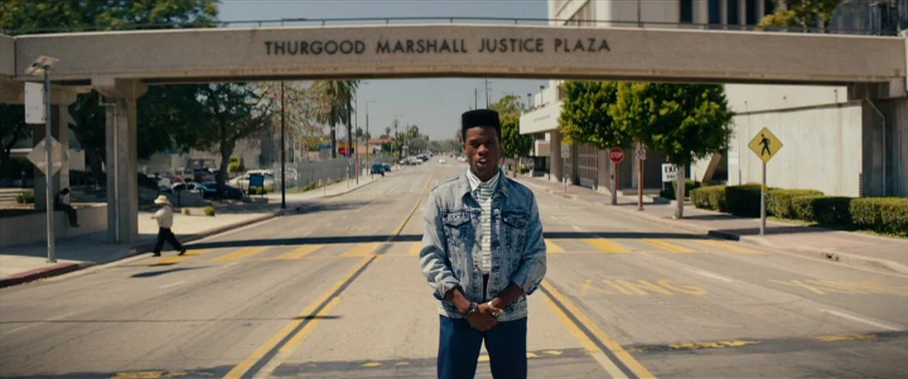 Dope (2015) movie still: fullbody — A man stands on a wide suburban street beneath an elevated concrete walkway. A sign on the walkway reads…