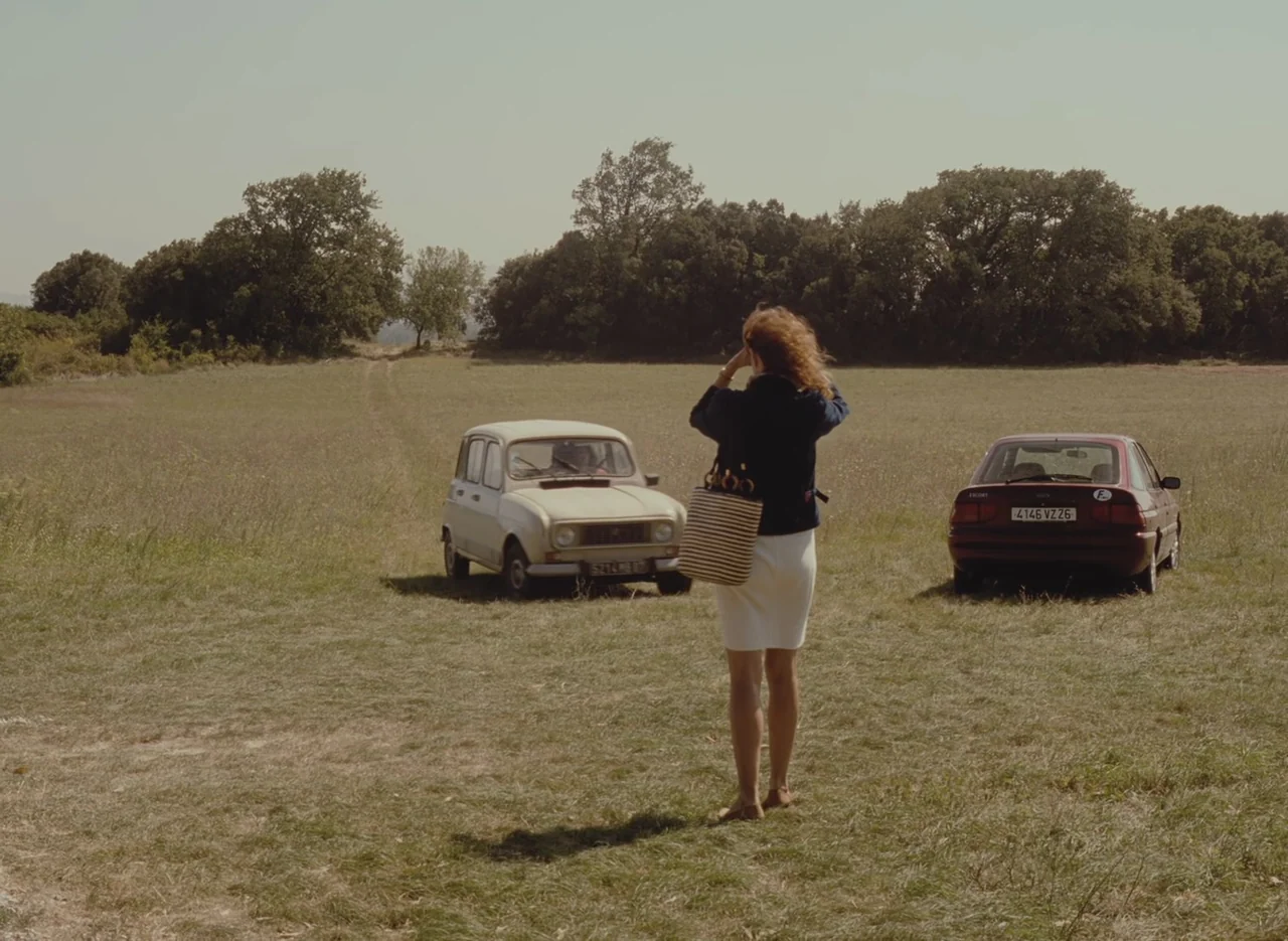 Autumn Tale (1998) movie still: fullbody — An open grassy field contains two parked vintage cars with a tree line beyond. A woman stands in the…
