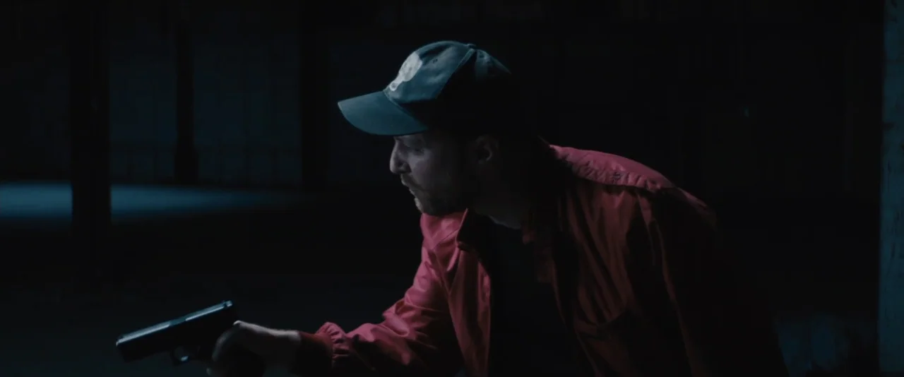 Stalker (2020) movie still: medium — A dark industrial space with vast shadows and indistinct architectural lines. A man in a red jacket crouches…