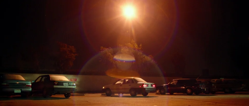 White Bird in a Blizzard (2014) movie still: wide — An asphalt parking lot at night features several cars parked in a row. An intense overhead…