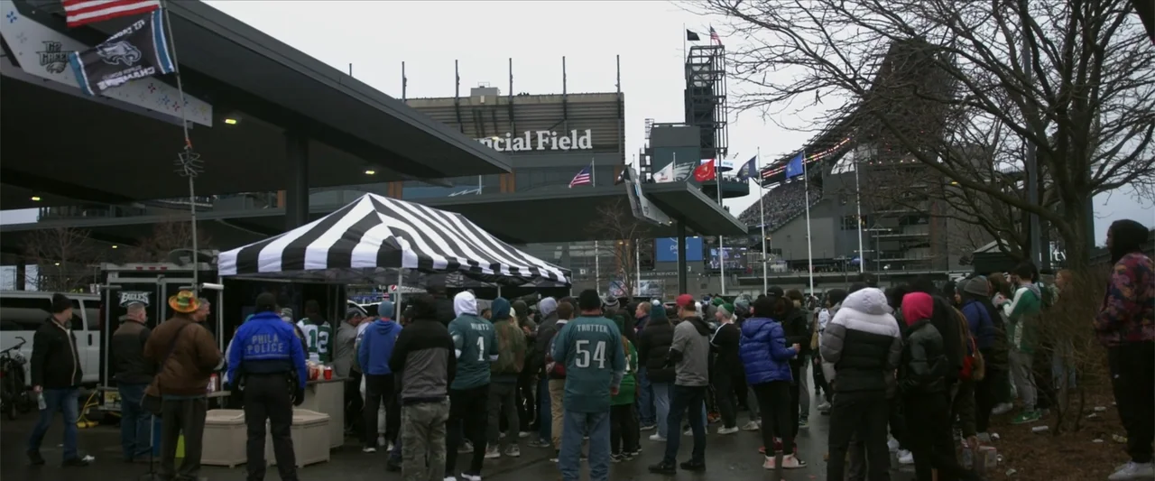 Kelce (2023) movie still: wide — A crowd of people wearing team jerseys stands in a paved outdoor plaza. They are positioned before a large stadium…
