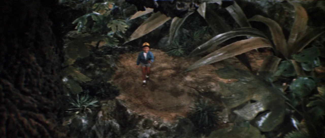 All Monsters Attack (1969) movie still: wide — A dense jungle clearing surrounded by oversized tropical leaves and dark, gnarled tree trunks. A…