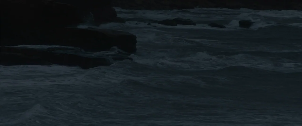 A Small Fortune (2021) movie still: establishing — A dark, rocky shoreline meets a turbulent sea during a low-light night setting. Waves break…