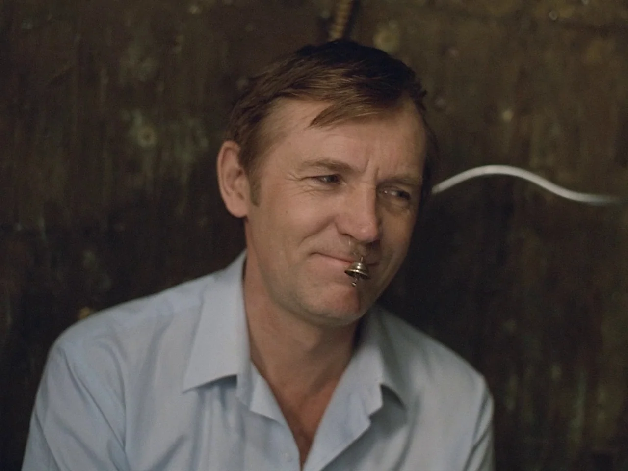 Kin-dza-dza! (1986) movie still: closeup — A man stands against a dark, textured wall surface in a dim room. The subject holds a small metal bell…