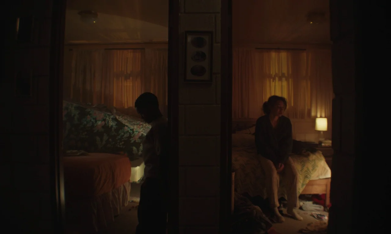 We Grown Now (2023) movie still: wide — A split composition shows two separate sleeping areas divided by a vertical concrete pillar. A young man…