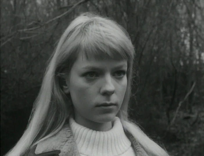Les Carabiniers (1963) movie still: closeup — A dense, leafless forest background frames a woman in a white turtleneck sweater. She looks slightly…