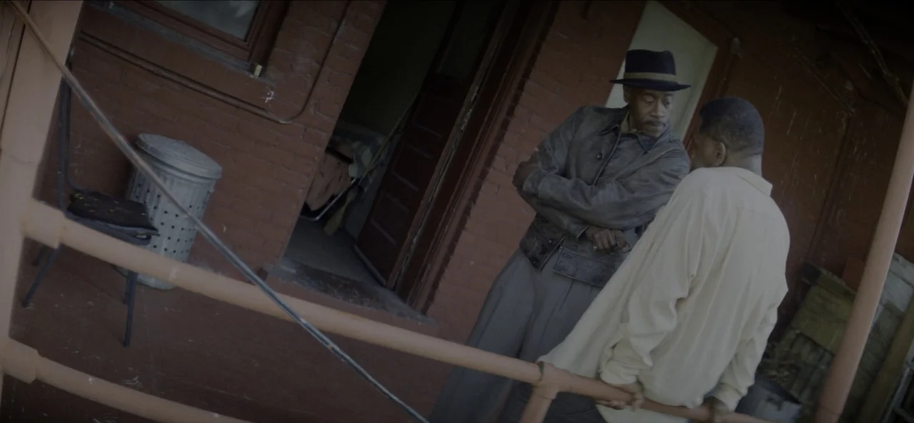 No Sudden Move (2021) movie still: medium — Two men stand on a weathered brick porch outside a simple building. The older man wears a fedora while…