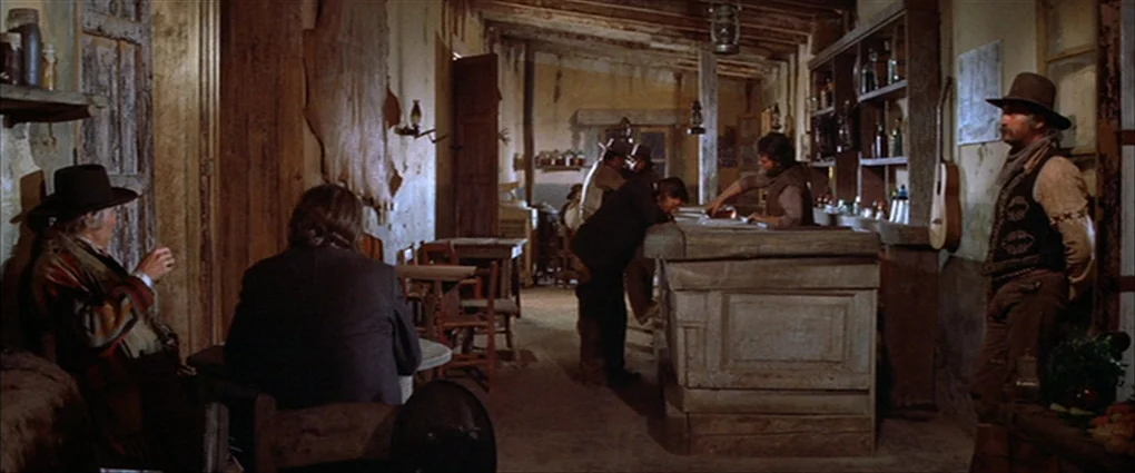 Pat Garrett & Billy The Kid (1973) movie still: wide — A rustic wood-paneled interior features a long bar counter and sparse wooden tables. Several…