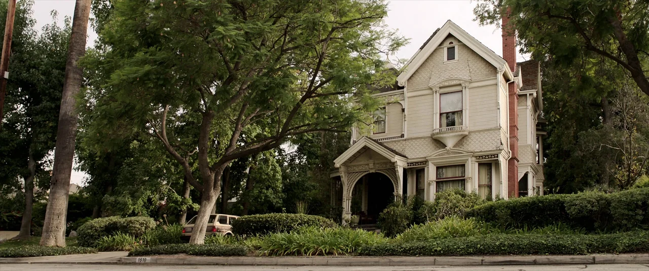 Malignant (2021) movie still: wide — A two-story Victorian house with light siding sits behind lush green trees. A vintage beige station wagon is…