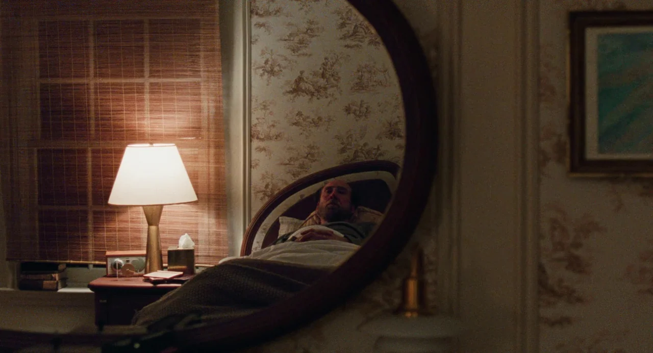 Dream Scenario (2023) movie still: medium — A bedroom features floral wallpaper and a lamp on a bedside nightstand. A man lies asleep in a bed…