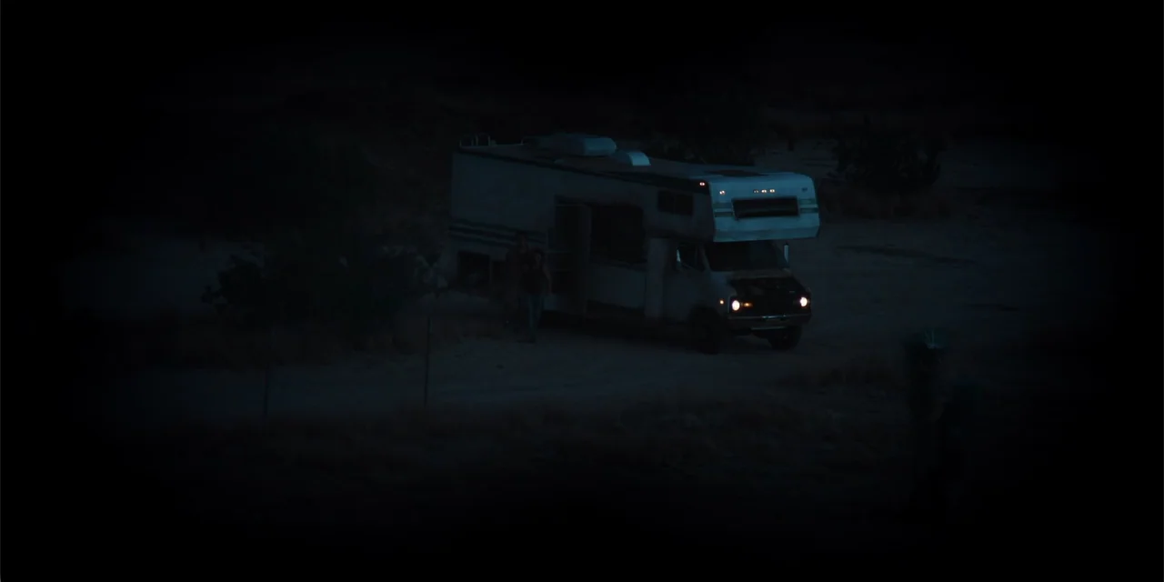Fall (2022) movie still: wide — A worn motorhome is parked on a barren, dark, desert patch at night. The vehicle's headlights cast a focused beam…