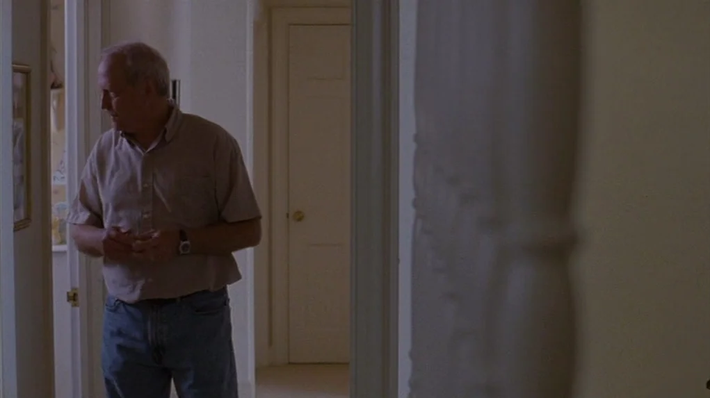 Junebug (2005) movie still: medium — A dim domestic hallway with white walls and closed doors on either side. An older man stands in the corridor…