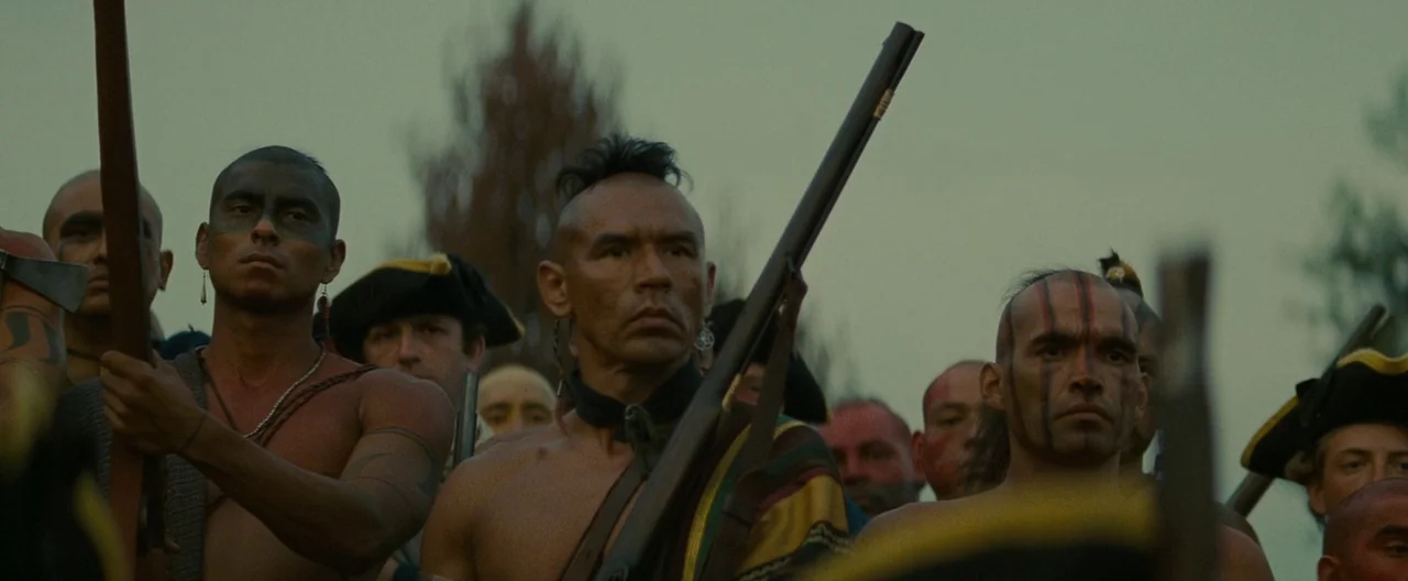 The Last of the Mohicans (1992) movie still: medium — A group of men with tribal markings and muskets gather in an open field. The overcast sky…