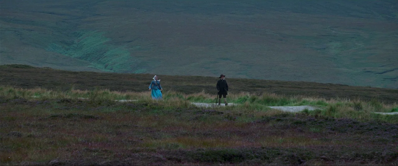 The Wonder (2022) movie still: wide — A vast, rolling green moorland features distant hills and a small dirt track. A man and a woman in…