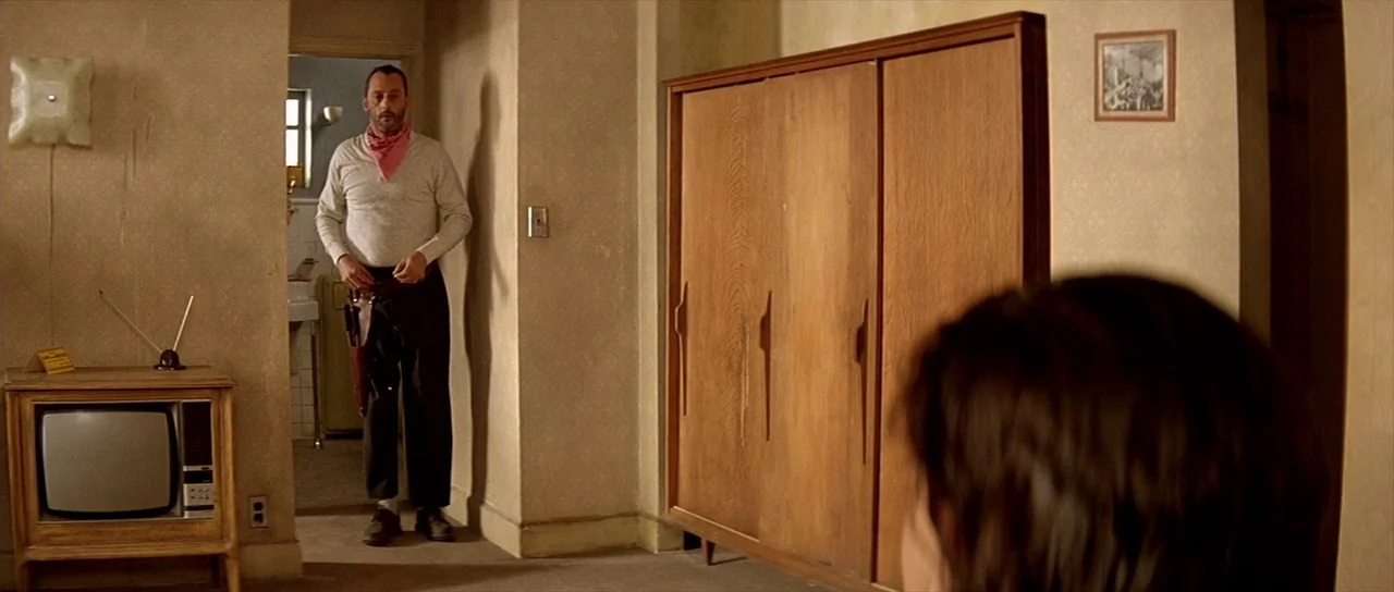 Léon: The Professional movie still: medium — A sparse interior with textured beige wallpaper, a large wooden armoire, and vintage television. A man…
