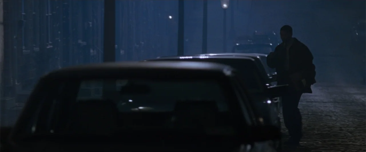 Fallen (2004) movie still: wide — A dark, wet cobblestone lane is lined with parked cars under dim street lighting. A solitary man in a dark jacket…