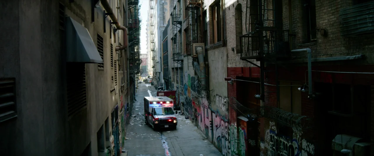 Ambulance (2022) movie still: wide — A narrow city alley features brick buildings, fire escapes, graffiti, and piles of trash. An ambulance with…