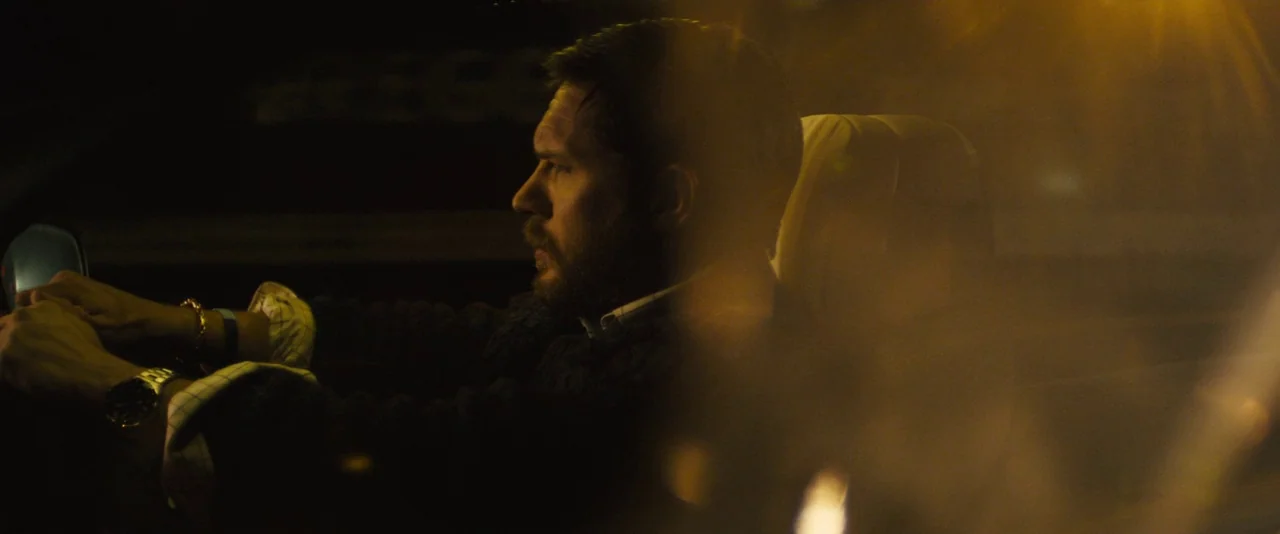 Locke (2013) movie still: medium — A man drives a car at night with amber light spilling through the window. The interior is dark and the man holds…