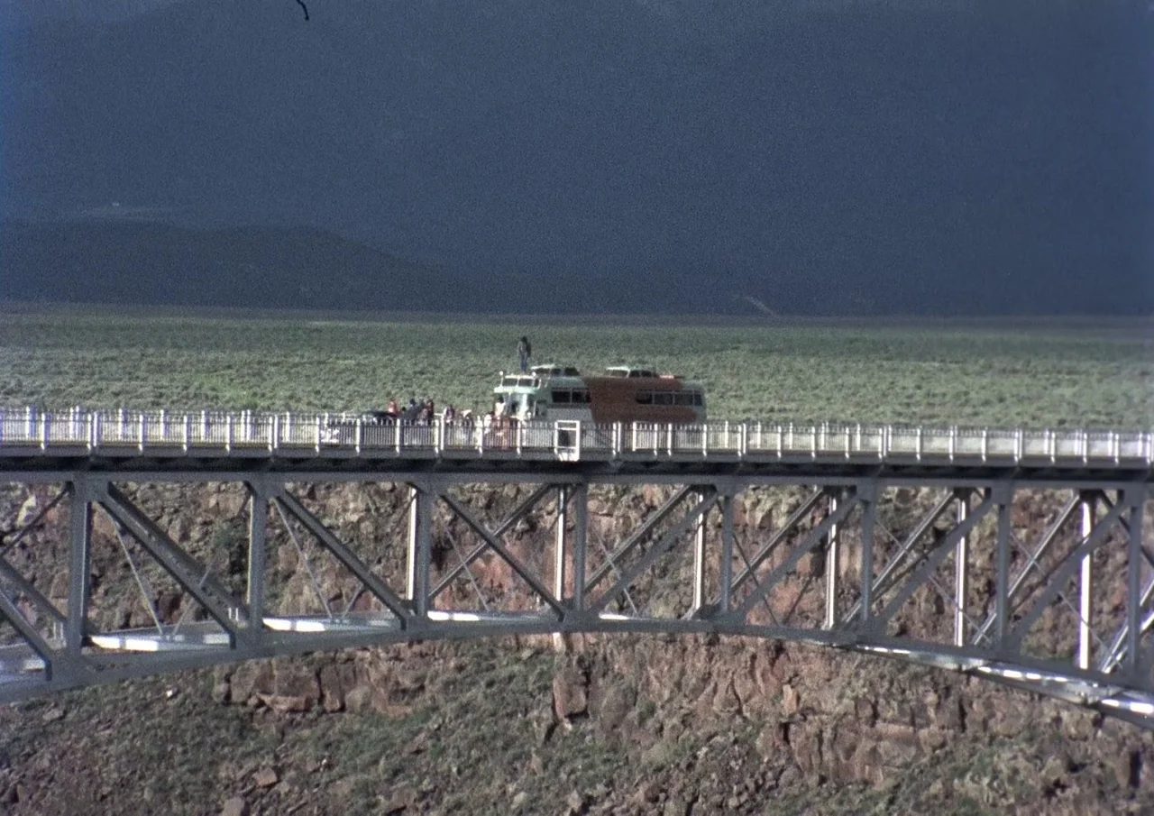 Human Highway (1982) movie still: wide — A long, industrial steel bridge spans a wide rocky gorge under a dark sky. A passenger bus sits stationary…