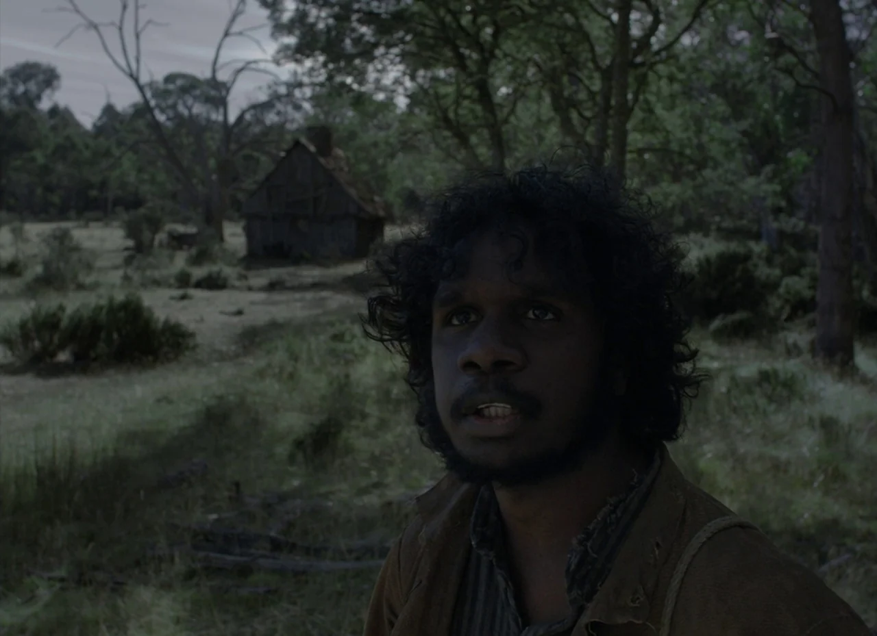 The Nightingale (2018) movie still: medium — A man stands in a sparse woodland area under a grey, overcast sky. A small, weathered wooden cabin is…