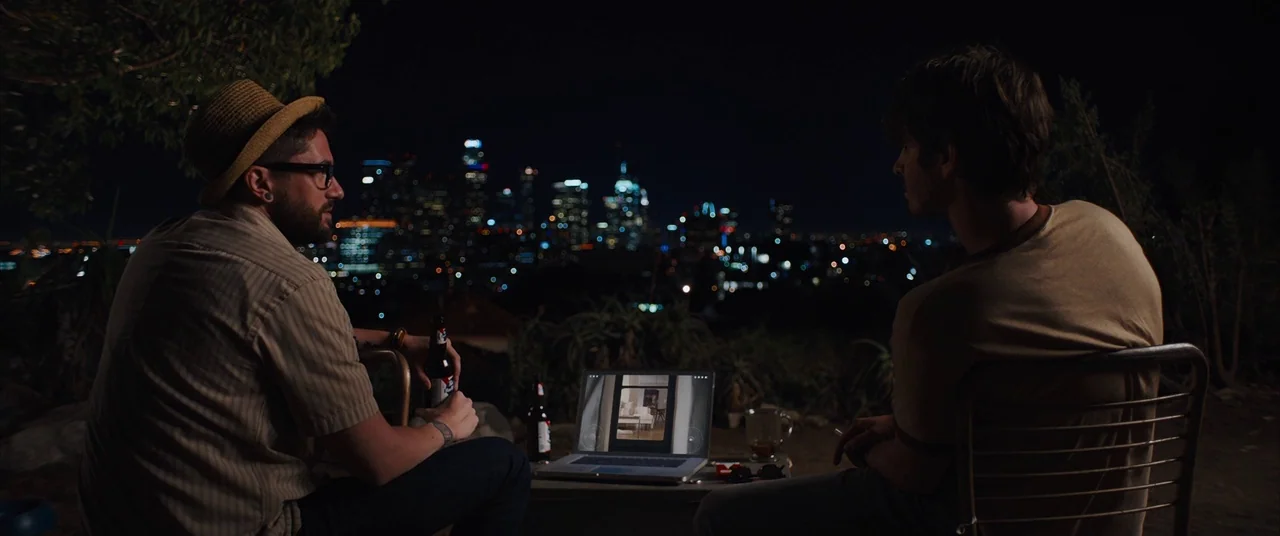 Under The Silver Lake (2018) movie still: wide — Two men sit at a table overlooking a blurred Los Angeles city skyline at night. They watch a laptop…