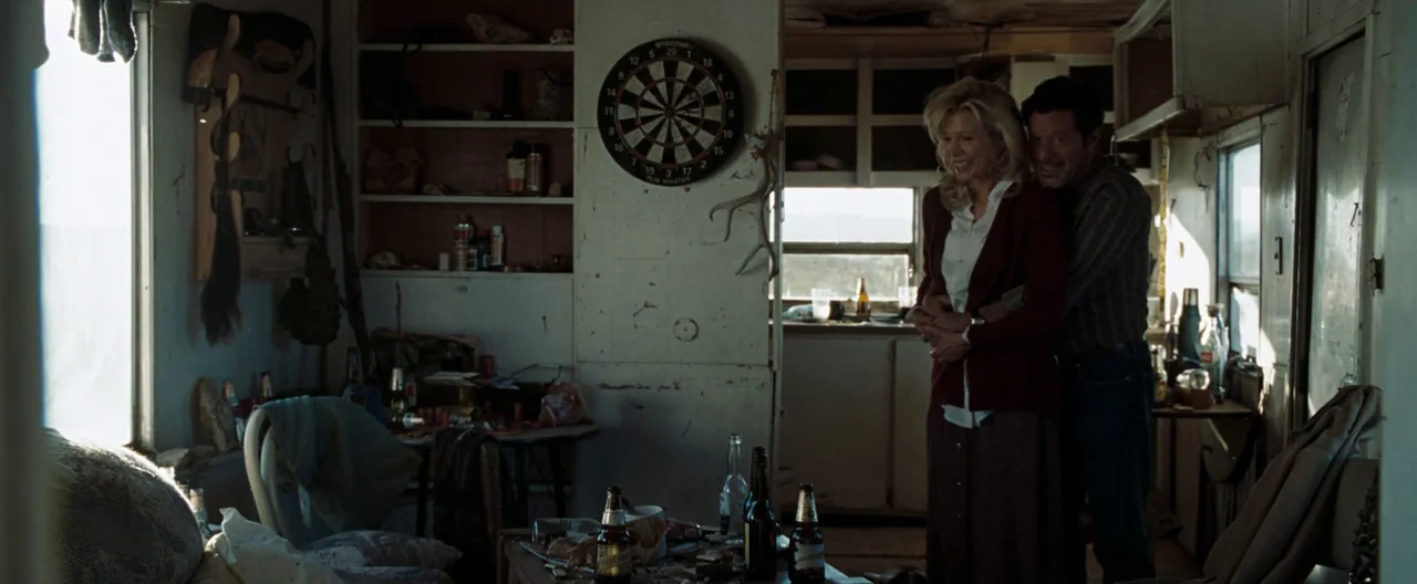 The Burning Plain (2008) movie still: medium — A cluttered, rustic mobile home interior features a dartboard on the wall and bottles. A man and…