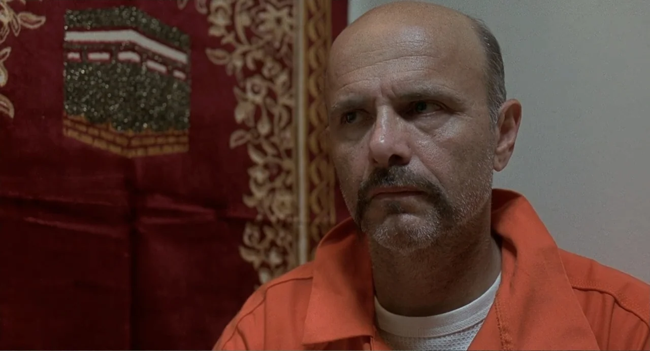 Wedding Daze (2006) movie still: medium — A man wearing an orange jumpsuit sits in front of a red fabric tapestry. He gazes off-camera with a…