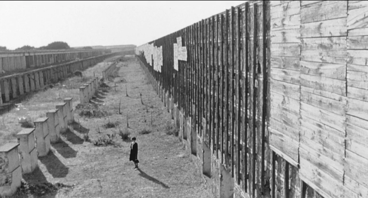 8 1/2 (1963) movie still: wide — A vast, dry field stretches alongside a long, weathered wooden fence with tattered posters. A lone woman stands…