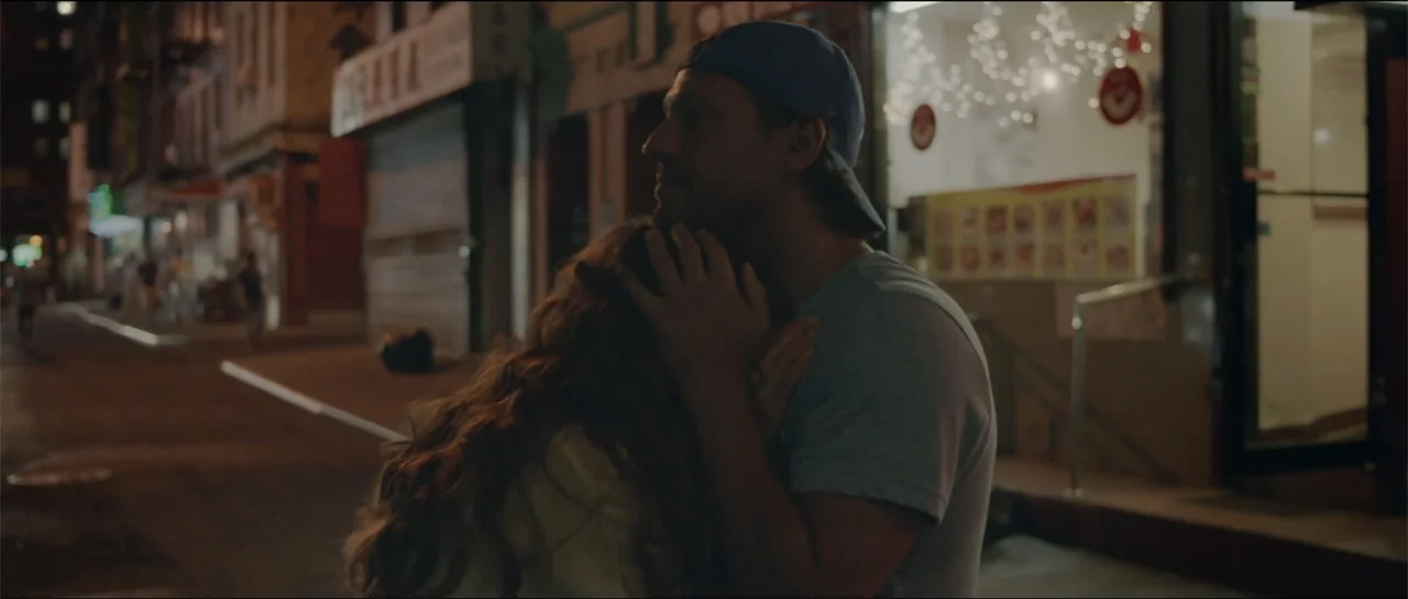 All She Wrote (2018) movie still: medium — A man and woman stand on a dimly lit urban sidewalk at night. The man holds the woman in a close embrace…