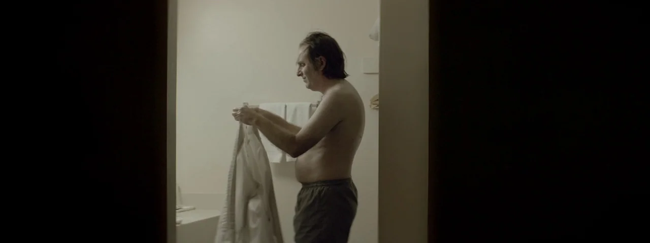 Entertainment (2015) movie still: medium — A spare, dimly lit room with plain light-colored walls and white hanging towels. A middle-aged man stands…