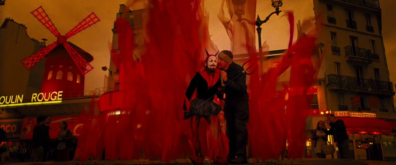 Micmacs (2009) movie still: fullbody — The exterior of the Moulin Rouge at night features a bright red illuminated windmill and signage. A man in a…