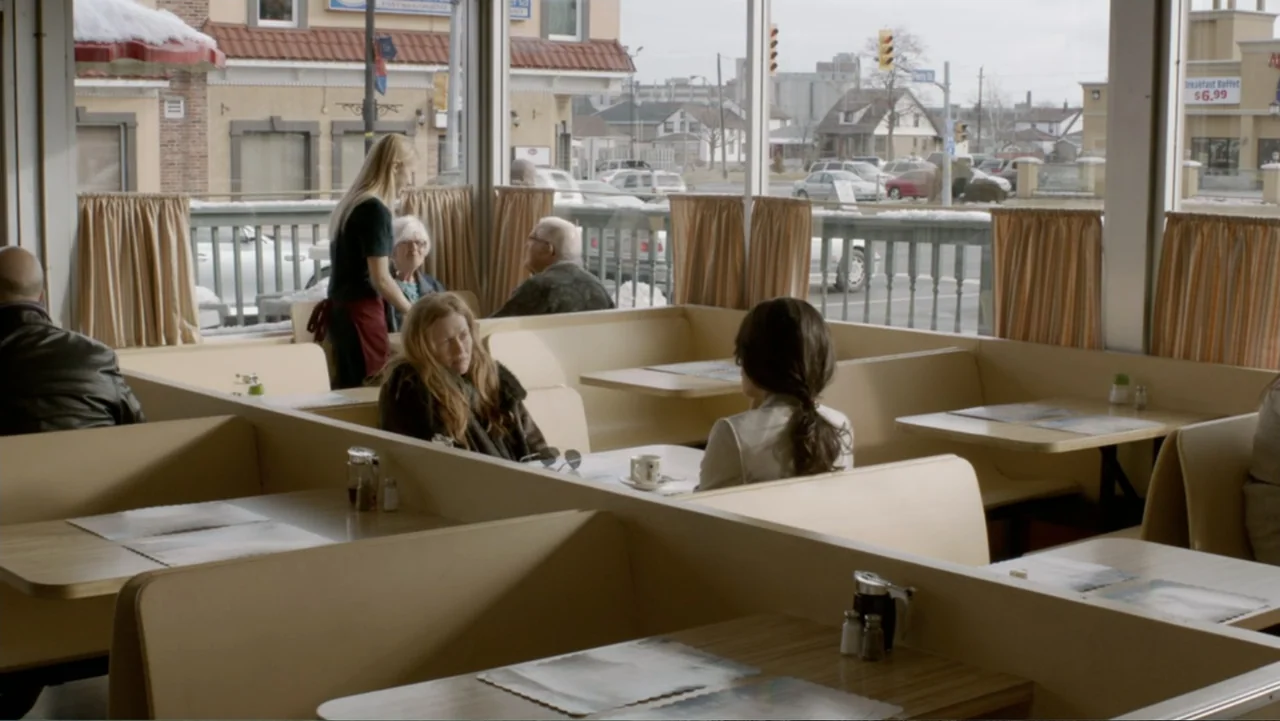 The Captive (2014) movie still: medium — A classic diner interior with beige booths and large windows overlooking a street. Two women sit at a table…