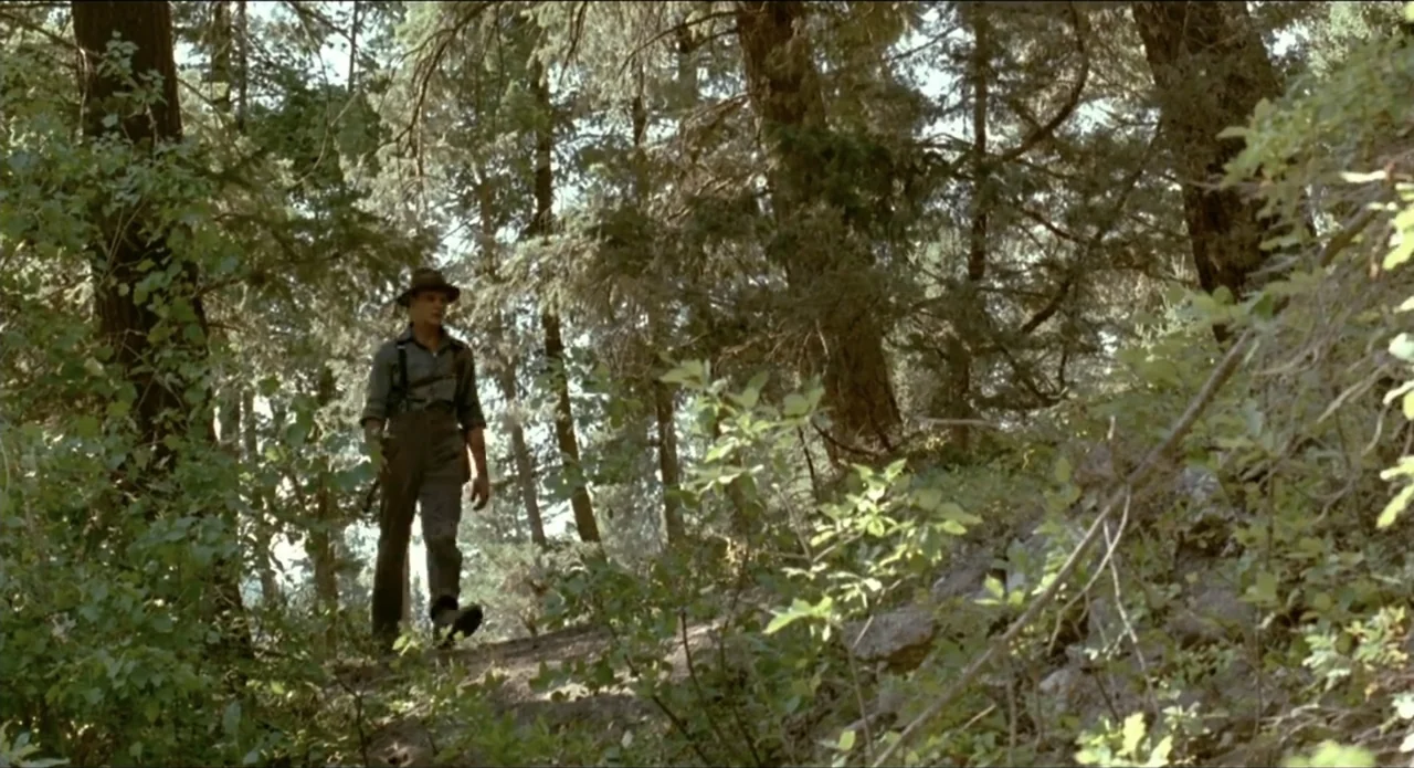 A River Runs Through It (1992) movie still: fullbody — A dense forest path filled with coniferous trees and uneven dirt ground. A man wearing a hat…