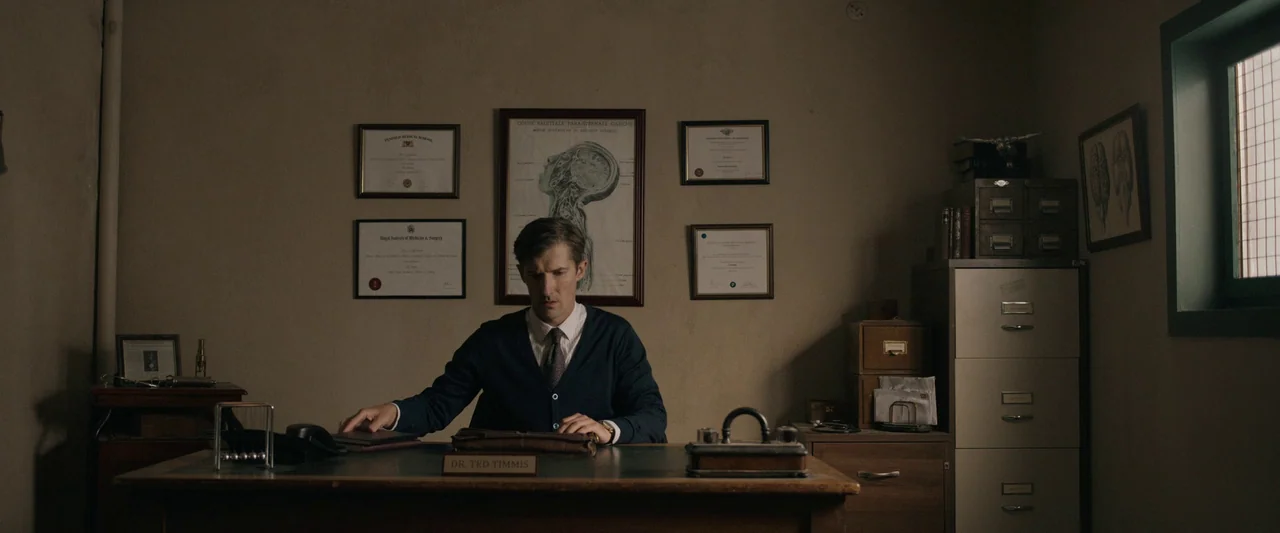 Oddity (2024) movie still: medium — A 1950s doctor's office contains a wooden desk with medical certificates on the wall. A man in a dark blue…