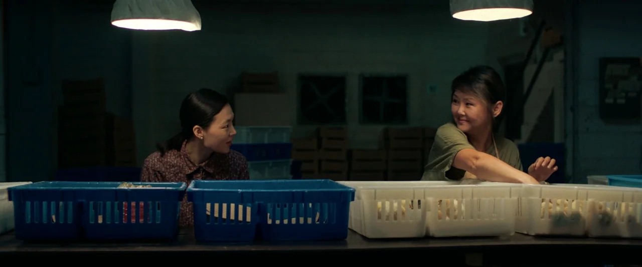 Minari (2020) movie still: medium — A dimly lit industrial room contains rows of plastic bins on a long table. Two women work alongside each other…