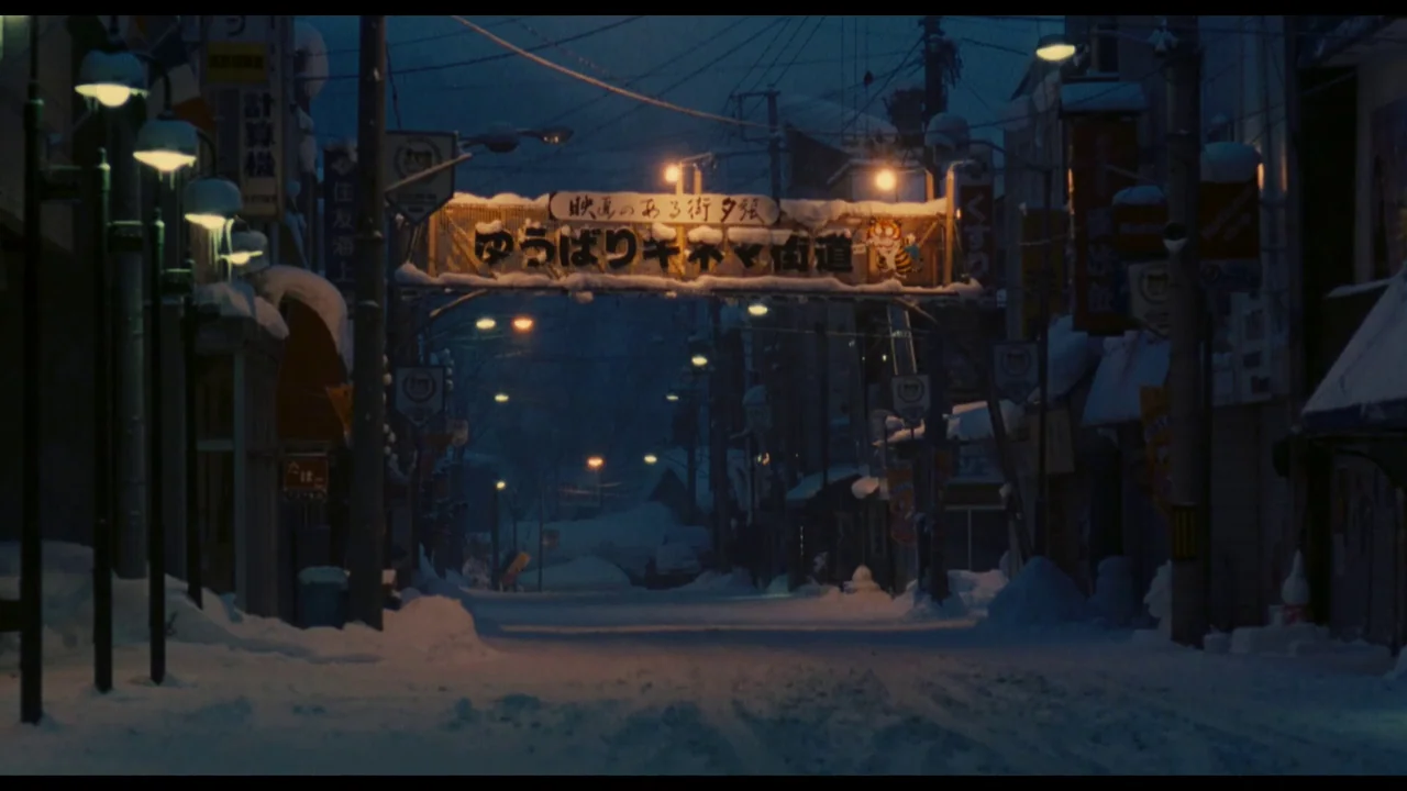 Millennium Mambo (2001) movie still: establishing — A quiet, snow-covered street in Yubari at night under a blue twilight sky. An arched sign…