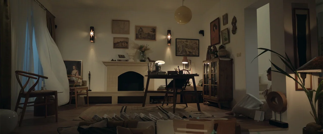Blue Cave (2024) movie still: wide — A living room with a fireplace, a writing desk, and assorted framed wall art. A woman sits on the wooden floor…