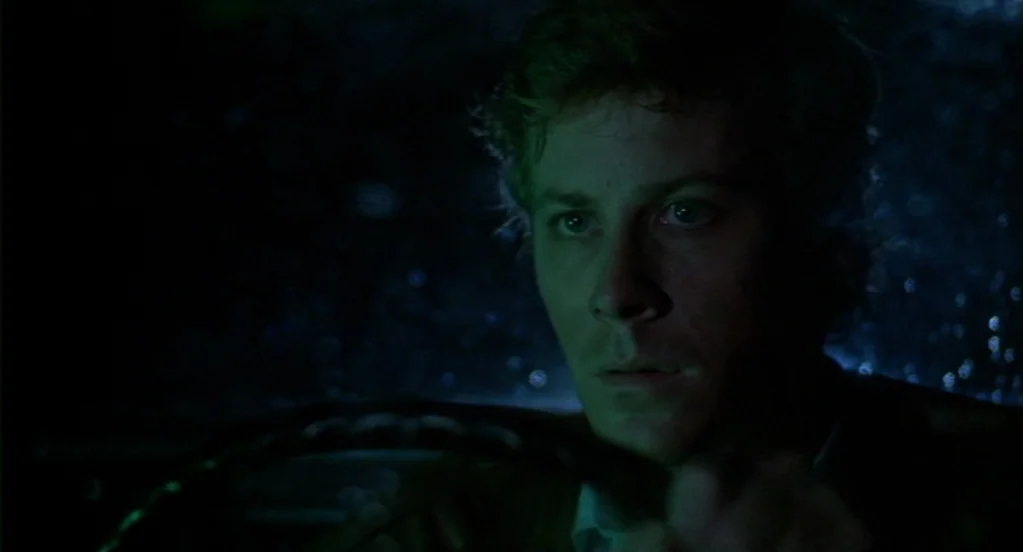 Cat People (1982) movie still: closeup — Inside a car cabin at night with rain visible against the window glass. A young man grips the steering…