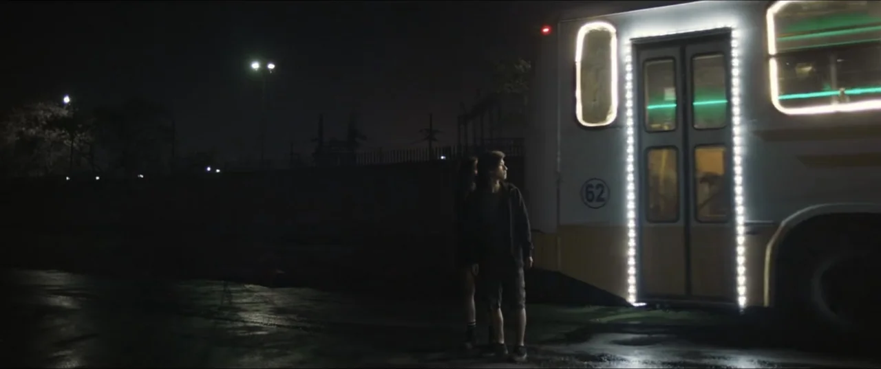 Death to Summer (2021) movie still: wide — A nighttime bus stop features a large bus with illuminated door frames parked nearby. Two teenagers wait…