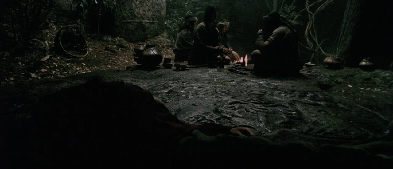 Renegade (2004) movie still: wide — A dark clearing features a large, flat, carved stone slab illuminated by fire. People sit around a flickering…