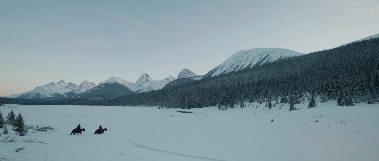 The Revenant (2015) movie still: wide — A vast snow-covered valley sits beneath a range of towering, rugged, snow-capped mountains. Two riders on…
