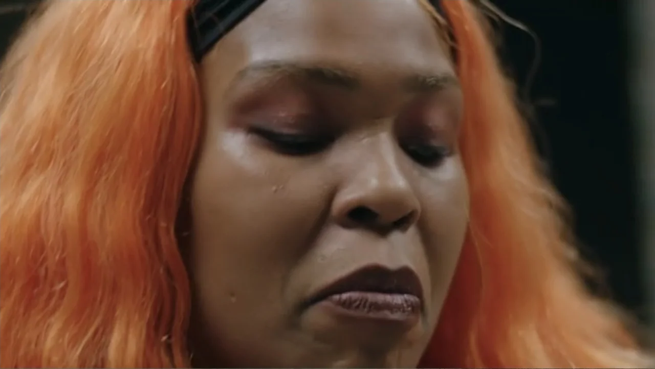 The African Desperate (2022) movie still: closeup — A close-up view shows a woman with bright orange hair inside a room. The subject maintains a…