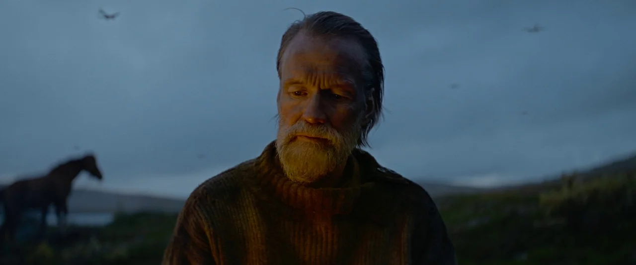 Sisu (2022) movie still: medium — A man stands on a desolate hillside at dusk beneath a cloudy sky. The man looks down toward the warmth of a fire…