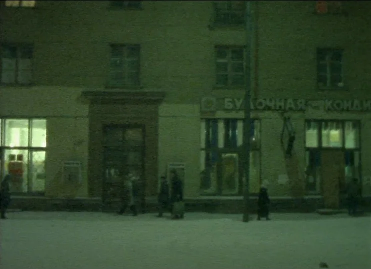From The East (1993) movie still: wide — A snow-covered street in front of a multi-story building at night. Several blurry figures move along the…