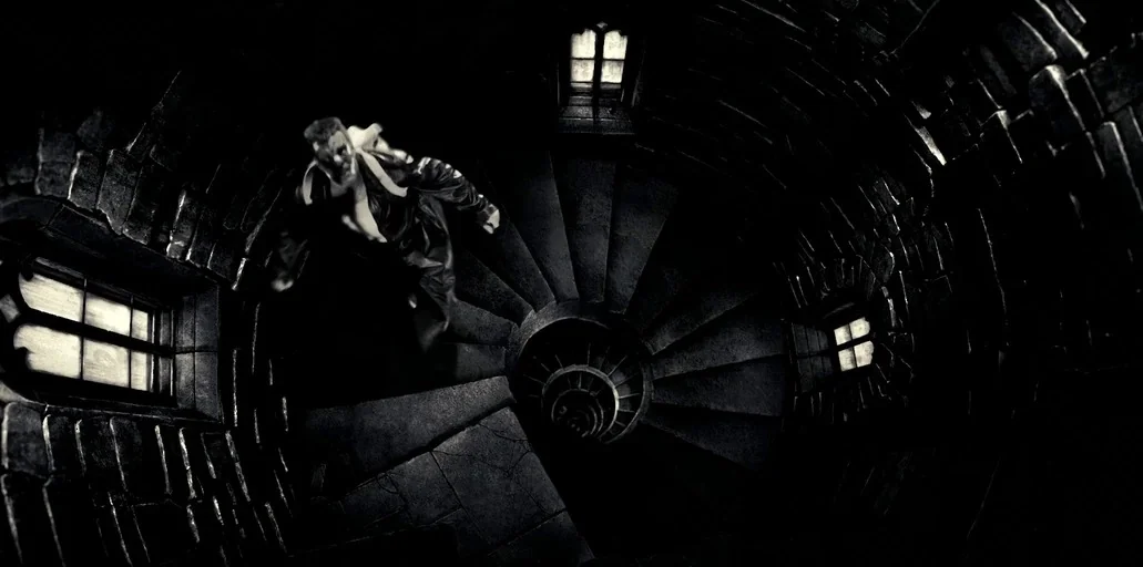 Sin City (2005) movie still: wide — A dark stone spiral tower staircase features small, glowing rectangular windows. A man moves quickly up the…