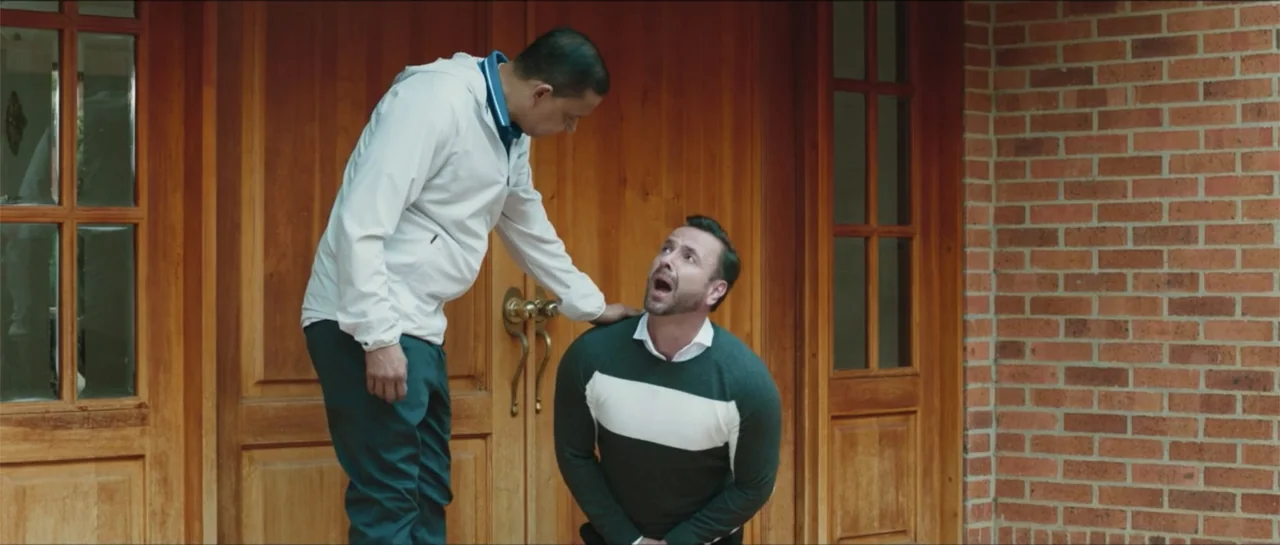 Loving is Losing (2019) movie still: medium — A house entrance features a large wooden double door and a brick wall. A man stands over another man…